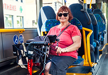 Frau mit rotem T-Shirt und schwarzem Rock sitzt in einem Bus auf einem blauen Sitz neben einem Rollator mit schwarzer Tasche.