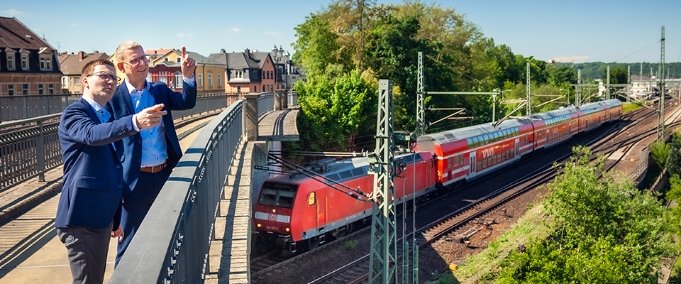 Zwei Männer stehen auf einer Brücke und zeigen auf eine rote S-Bahn, die auf Gleisen neben der Brücke fährt.